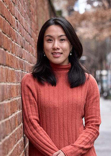A woman in an orange sweater smiles leaning against a brick wall on a city street